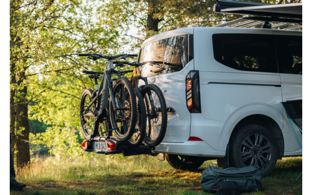 Weißer Camper-Van auf Wiese unter Bäumen; drei Fahrräder am Heck auf Fahrradträger; Reisetasche neben dem Fahrzeug