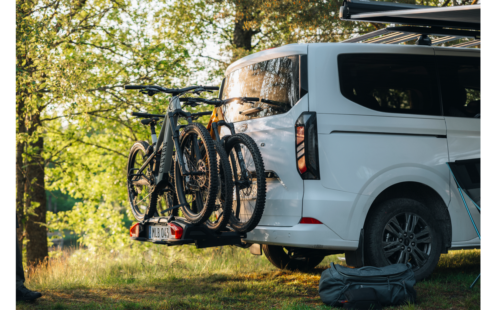 Weißer Camper-Van auf Wiese unter Bäumen; drei Fahrräder am Heck auf Fahrradträger; Reisetasche neben dem Fahrzeug