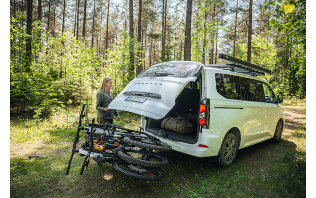 Weißer Van mit offenem Heck auf Waldlichtung, eine Person steht am Heck, zwei Mountainbikes an Heckträger, Gepäck im Laderaum sichtbar, Dachträger mit langem Gepäck
