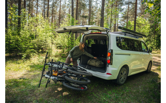 weißer Van mit geöffneter Heckklappe auf Waldlichtung, eine Person beugt sich in den Kofferraum, drei Mountainbikes liegen nebeneinander auf dem Boden, Bäume und Gras im Hintergrund