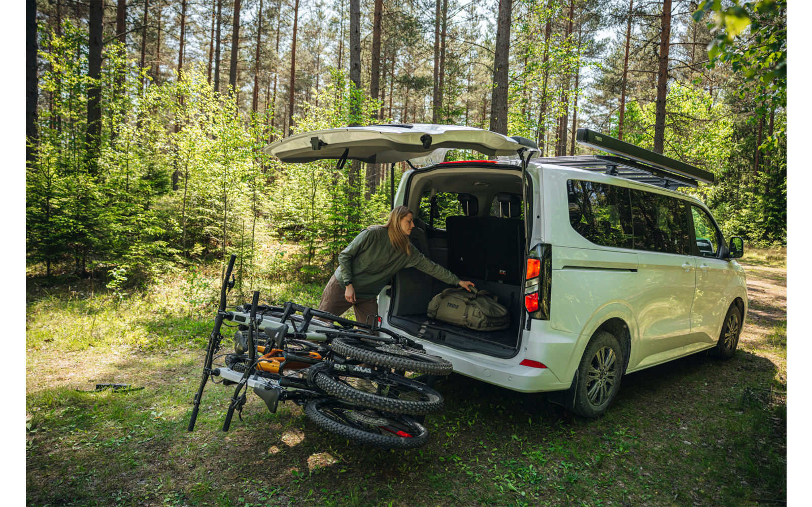 weißer Van mit geöffneter Heckklappe auf Waldlichtung, eine Person beugt sich in den Kofferraum, drei Mountainbikes liegen nebeneinander auf dem Boden, Bäume und Gras im Hintergrund