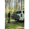 Zwei Personen neben einem weißen Van im Wald; eine Person mit Helm befestigt drei Fahrräder an einem Heckträger, hohe Nadelbäume und Gras im Hintergrund