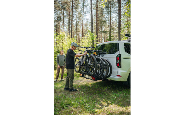 Zwei Personen neben einem weißen Van im Wald; eine Person mit Helm befestigt drei Fahrräder an einem Heckträger, hohe Nadelbäume und Gras im Hintergrund