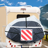 Vista posteriore di un camper chiaro con una ruota di scorta coperta da una copertura nera e grigio chiaro con una targa di segnalazione rossa e bianca a strisce diagonali; finestrino posteriore, fanali e targa visibili; lago, montagne e cielo azzurro con nuvole sullo sfondo