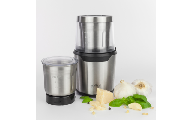 Gerät aus Edelstahl mit zwei abnehmbaren Behältern; davor Knoblauchknolle, Knoblauchzehen, Stück und Raspel Parmesan, Basilikumblätter, Pinienkerne und schwarze Pfefferkörner