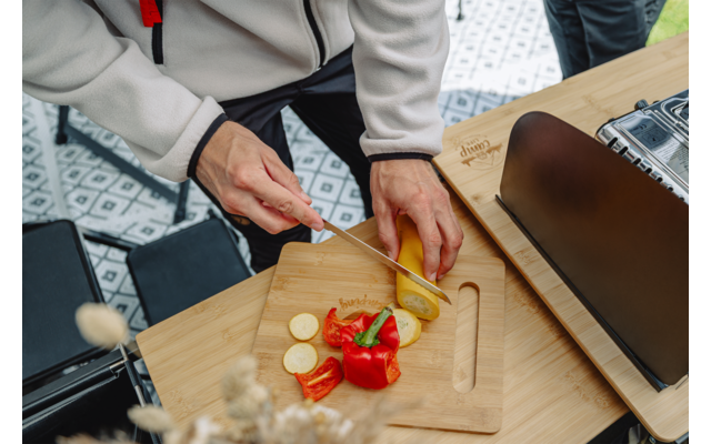 Hände schneiden eine gelbe Zucchini auf einem Holz-Schneidebrett; daneben geschnittene rote Paprika, ein Messer und ein schwarzes Gerät auf einem Holztisch