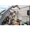 Wohnwagen mit ausgefahrener Markise; ein Mann sitzt an einem Campingtisch mit einer Schüssel Salat, Geschirr, Laterne und Thermosflasche; eine Frau steht in der Tür; zwei Campingstühle; grünes Feld und Himmel im Hintergrund