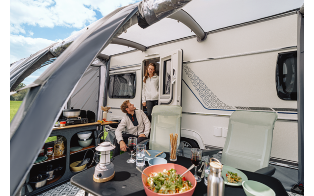 Wohnwagen mit ausgefahrener Markise; ein Mann sitzt an einem Campingtisch mit einer Schüssel Salat, Geschirr, Laterne und Thermosflasche; eine Frau steht in der Tür; zwei Campingstühle; grünes Feld und Himmel im Hintergrund