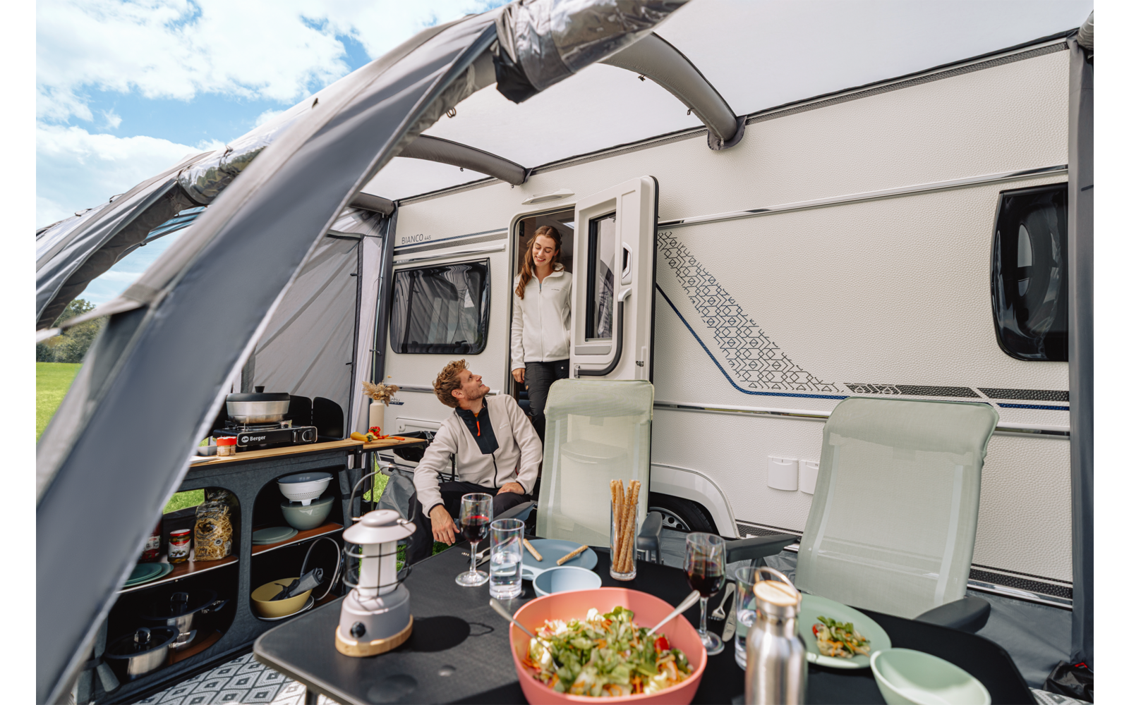 Wohnwagen mit ausgefahrener Markise; ein Mann sitzt an einem Campingtisch mit einer Schüssel Salat, Geschirr, Laterne und Thermosflasche; eine Frau steht in der Tür; zwei Campingstühle; grünes Feld und Himmel im Hintergrund
