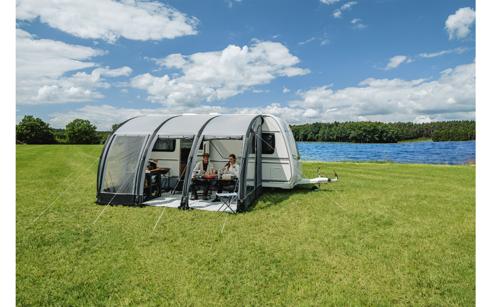 Wohnwagen mit Vorzelt, zwei Personen an einem Tisch, Wiese, See und bewölkter Himmel