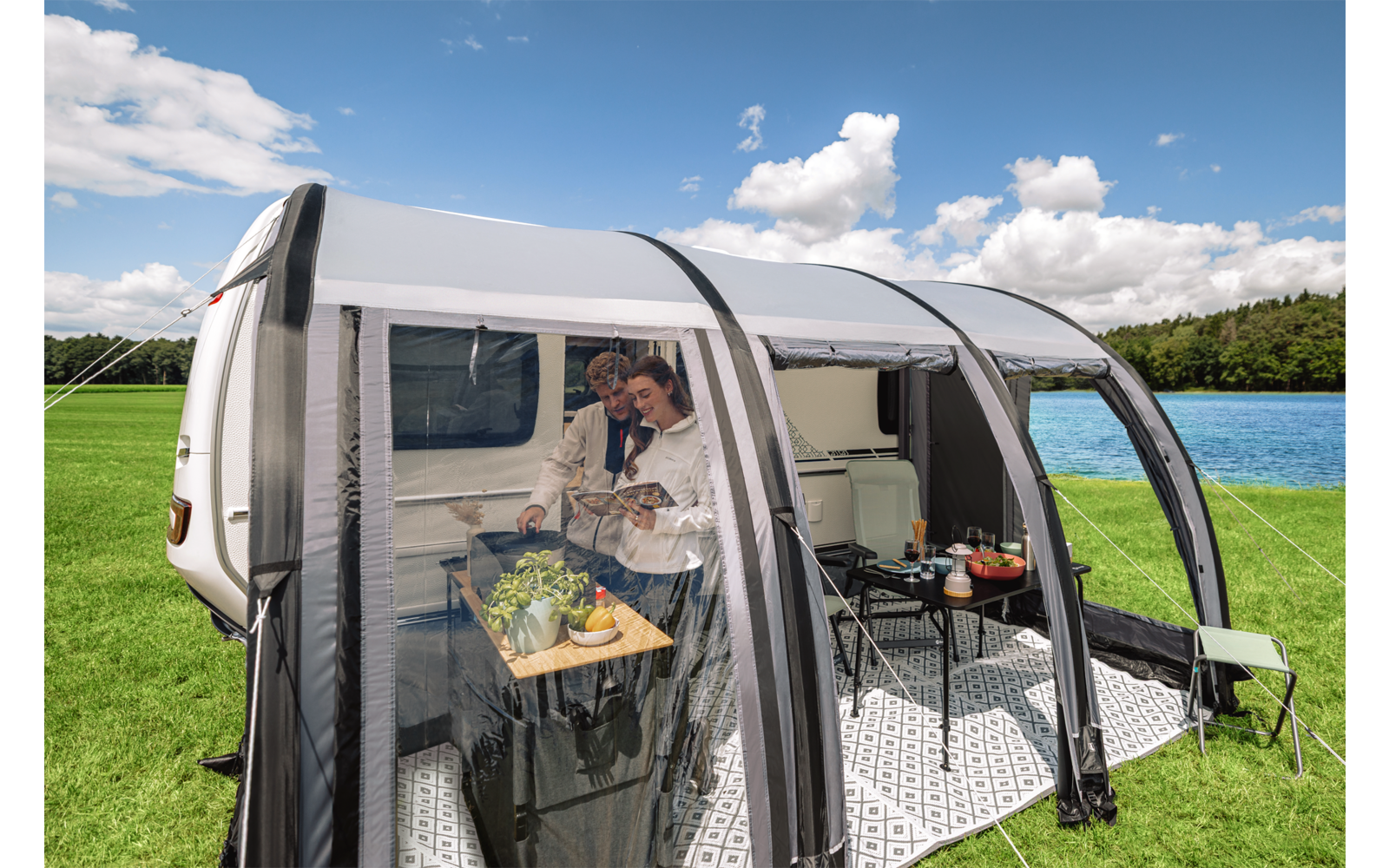 Weißer Wohnwagen mit offenem, gebogenem Vorzelt aus transparenten Planen auf einer Wiese neben einem See; im Vorzelt zwei Personen, ein Tisch mit Pflanzen und Lebensmitteln, Sitzgelegenheiten
