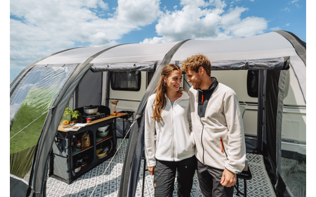 Zwei Personen stehen nebeneinander im Eingang eines Vorzelts an einem Wohnwagen; links ist eine Campingküche mit Utensilien sichtbar; darüber leicht bewölkter Himmel