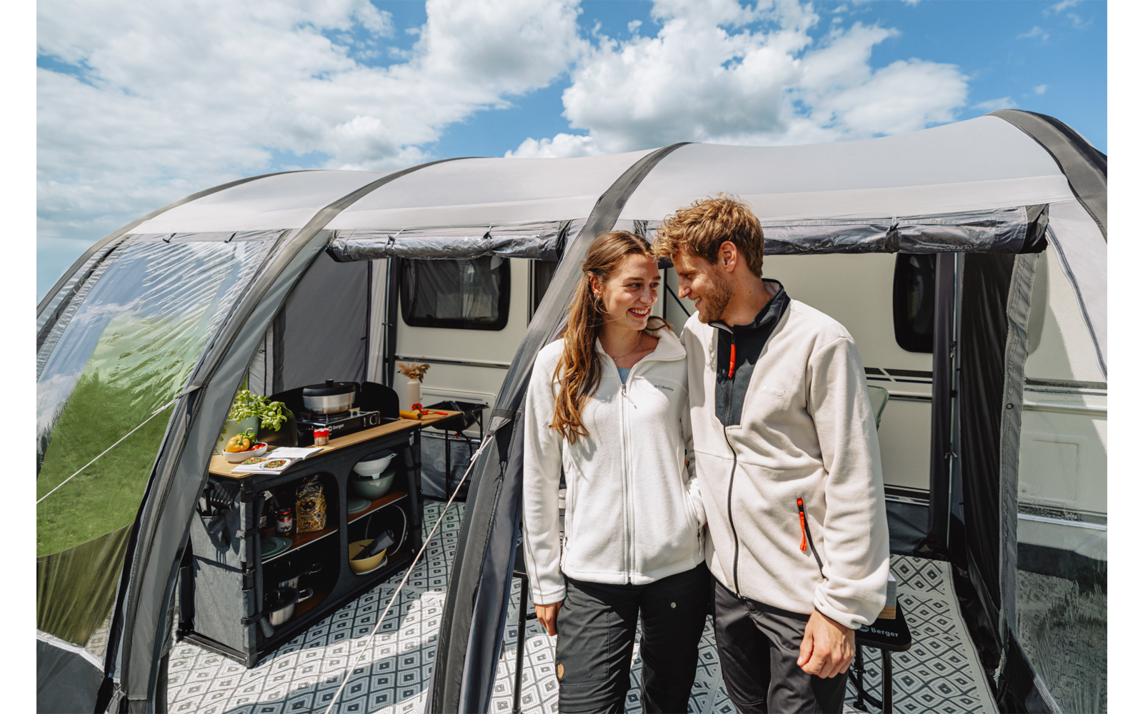 Zwei Personen stehen nebeneinander im Eingang eines Vorzelts an einem Wohnwagen; links ist eine Campingküche mit Utensilien sichtbar; darüber leicht bewölkter Himmel