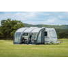 Weißer Wohnwagen mit grauem aufblasbarem Vorzelt auf einer Wiese, zwei Campingstühle und ein kleiner Tisch sichtbar, Bäume und bewölkter Himmel im Hintergrund