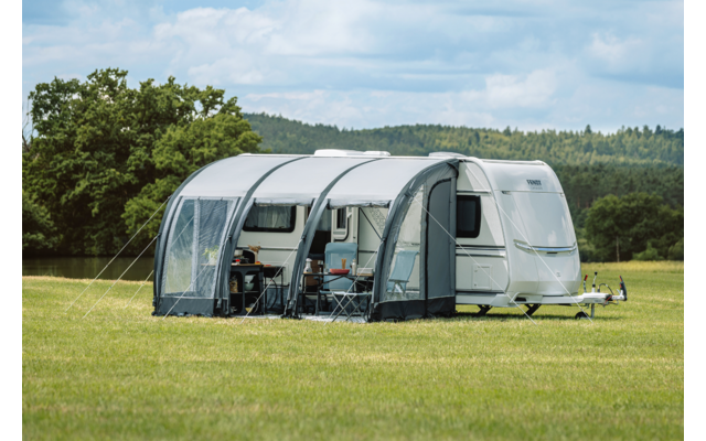 Weißer Wohnwagen mit grauem aufblasbarem Vorzelt auf einer Wiese, zwei Campingstühle und ein kleiner Tisch sichtbar, Bäume und bewölkter Himmel im Hintergrund