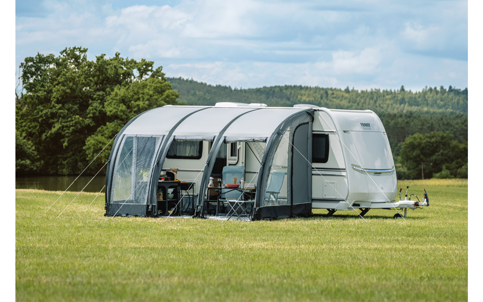 Weißer Wohnwagen mit grauem aufblasbarem Vorzelt auf einer Wiese, zwei Campingstühle und ein kleiner Tisch sichtbar, Bäume und bewölkter Himmel im Hintergrund