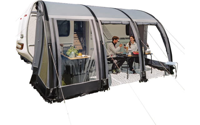 Caravane blanche avec auvent accolé sur une prairie verte au bord d’un lac ; deux personnes assises à une table dans l’auvent ; ciel bleu avec des nuages et une rangée d’arbres en arrière-plan