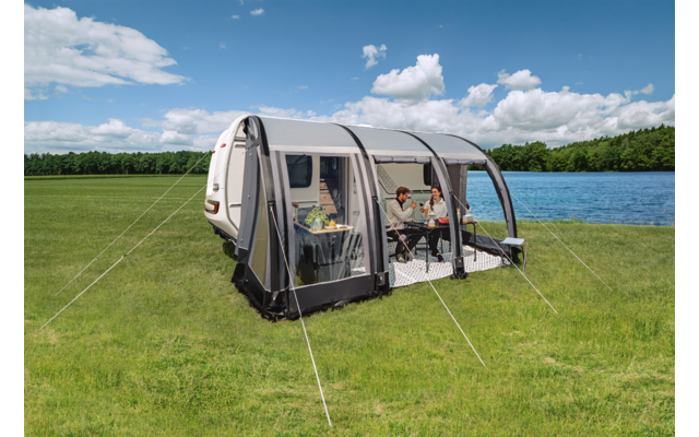 Caravan bianco con tenda annessa su un prato verde accanto a un lago; due persone sedute a un tavolo nella tenda; cielo azzurro con nuvole e una fila di alberi sullo sfondo