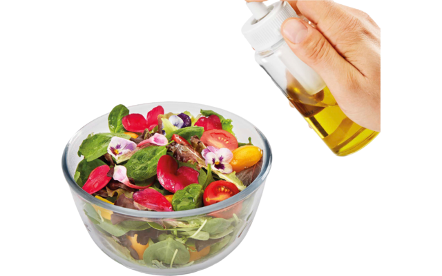 Glas-Schüssel mit gemischten Salatblättern, Radieschenscheiben, kleinen roten und gelben Tomaten sowie lila und rosa Blüten; eine Hand hält eine Glasflasche mit gelber Flüssigkeit über dem Salat
