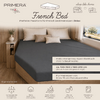 Donkergrijs hoeslaken op een French-bed in een camperinterieur, drie beige kussens bij het hoofdeinde, houten bedframe, klein nachtkastje met vaas en droogbloemen, raam met landschap, producttekst en logo's zichtbaar