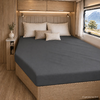 Tweepersoonsbed in een camper met donkergrijs hoeslaken, drie kussens, houten wandbekleding, nachtkastje met vaas en raam met uitzicht op een meer en bergen