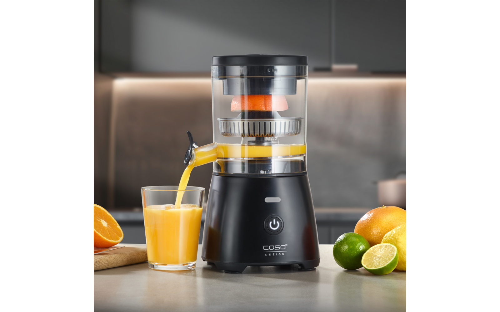 Extracteur de jus électrique noir sur le plan de travail de la cuisine, le jus coule du bec dans un verre, demi-orange sur une planche à gauche, oranges entières et citrons verts à droite