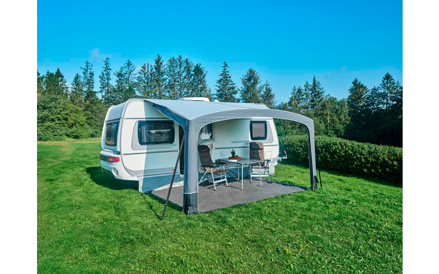 Weißer Wohnwagen mit grauem Vorzelt auf Gras, zwei Campingstühle und ein Tisch, Bäume und blauer Himmel im Hintergrund