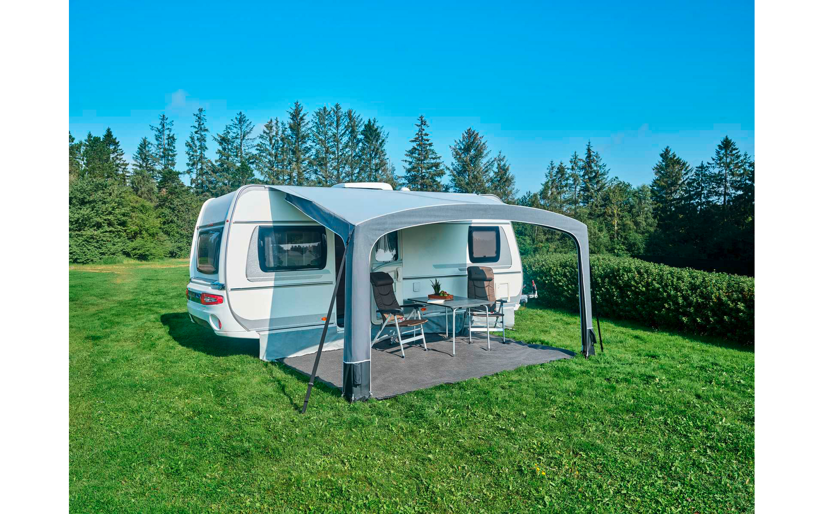Weißer Wohnwagen mit grauem Vorzelt auf Gras, zwei Campingstühle und ein Tisch, Bäume und blauer Himmel im Hintergrund