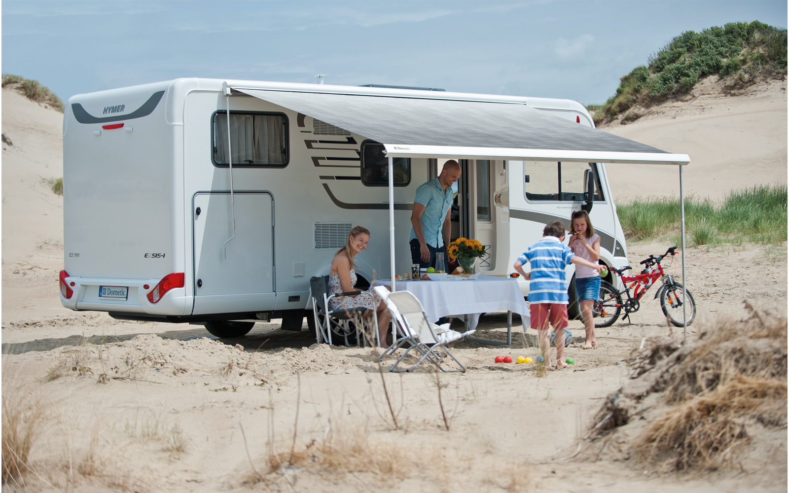 Camping-car avec auvent dans un paysage sablonneux, trois personnes assises et debout devant, un vélo appuyé contre le véhicule