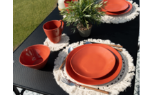 Une table dressée avec des assiettes orange, des tasses et des dessous de verre en crochet blanc sur une nappe noire, avec de l'herbe verte en arrière-plan