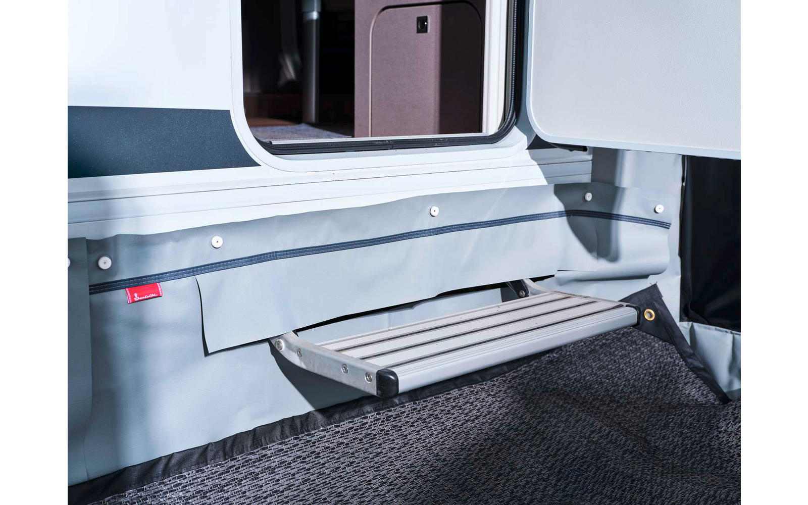 Marche pliante en aluminium devant une porte ouverte de caravane, jupe en tissu gris et paillasson foncé visibles