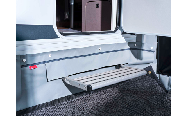 Marche pliante en aluminium devant une porte ouverte de caravane, jupe en tissu gris et paillasson foncé visibles