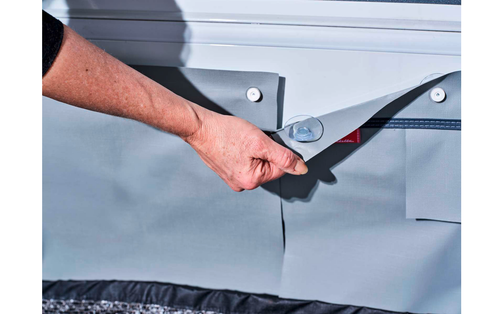 Main tire sur un anneau en plastique rond et soulève un rabat en tissu gris ; visible en dessous : détail triangulaire rouge, coutures grises et ourlet noir