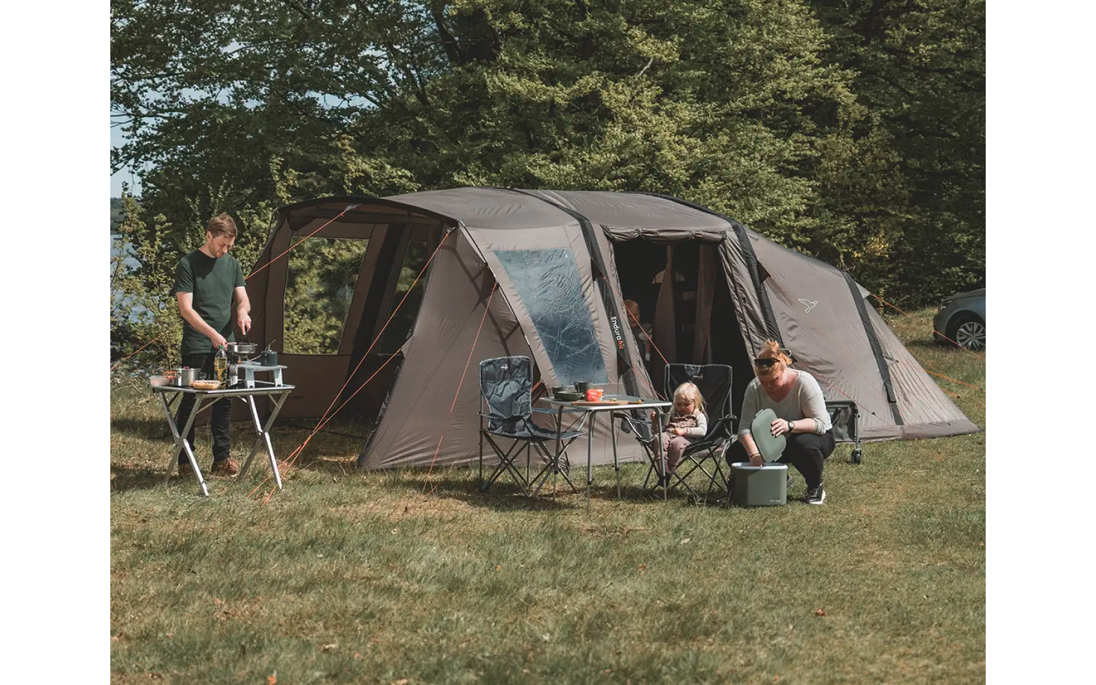 Großes graues Zelt auf einer Wiese mit vier Personen, Bäumen im Hintergrund