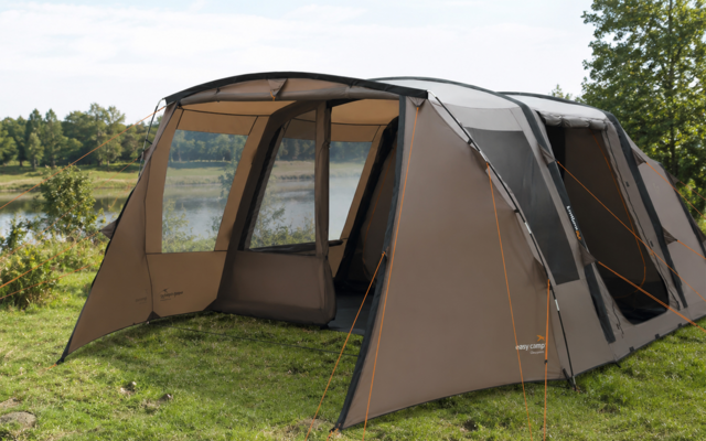 Large brown-gray tent with open front awning on grass by a lake, with trees and sky in the background