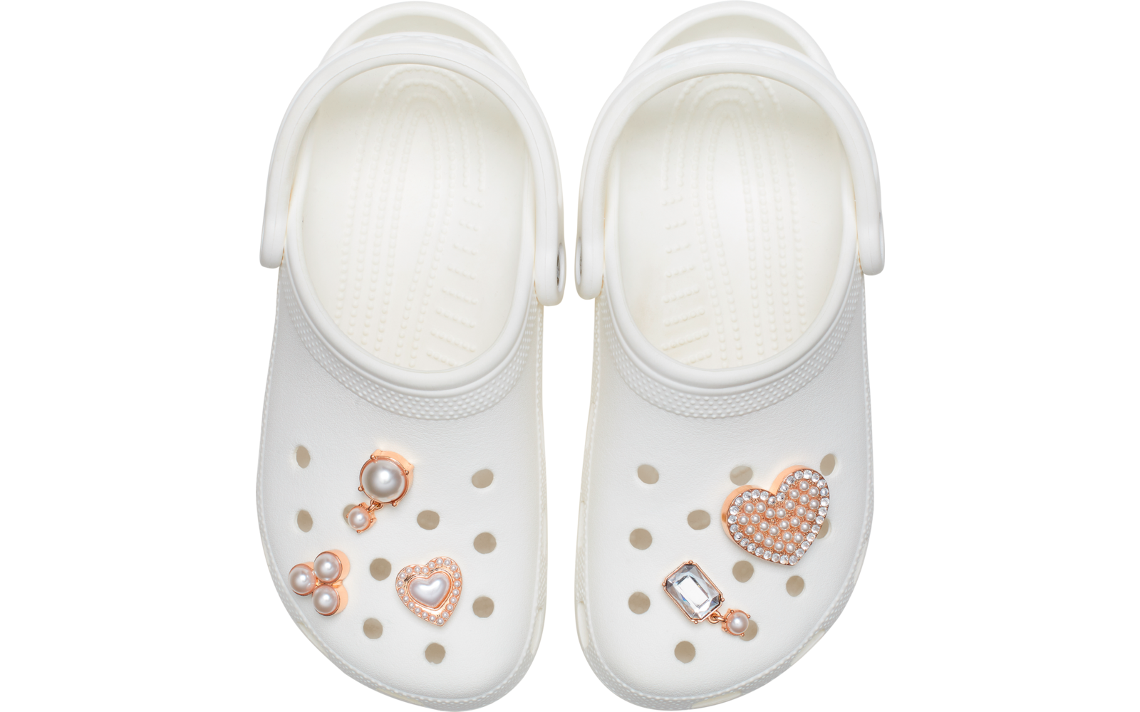 Dos zuecos blancos con agujeros, decorados con dijes: perlas, un corazón con pedrería y una gema rectangular, vista superior