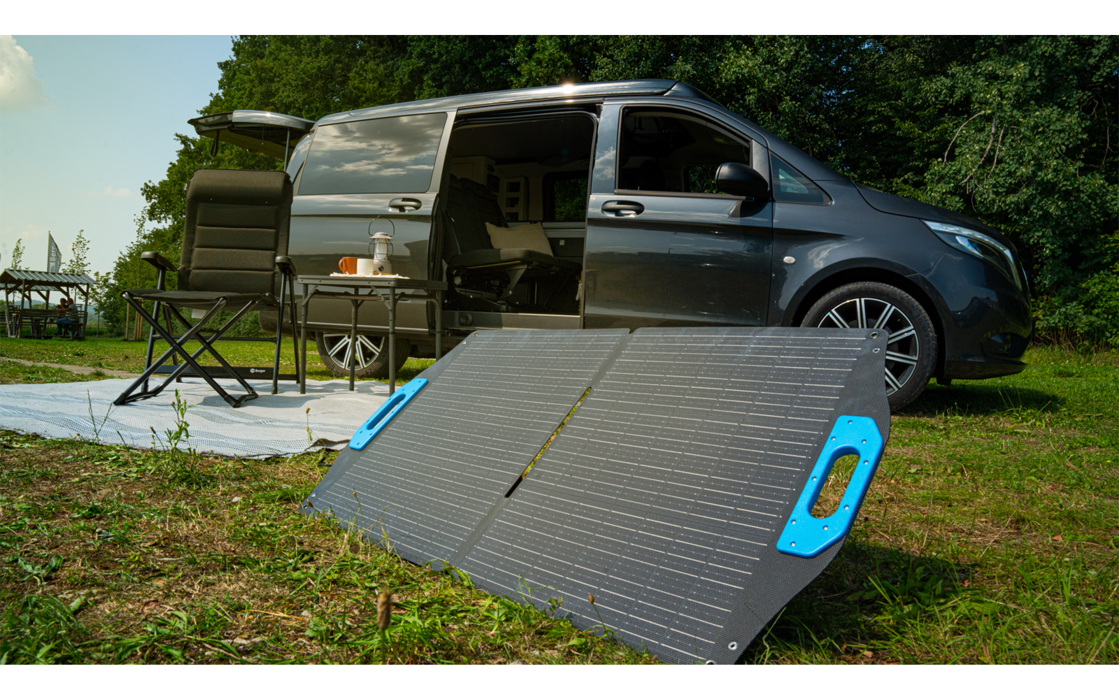 Panel solar portátil con asas azules sobre la hierba en primer plano; al fondo una furgoneta gris con puerta corredera abierta, una alfombra blanca, una silla plegable y árboles bajo un cielo azul