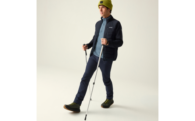 Person in Outdoor-Bekleidung mit grünem Strickmütze, dunkler Jacke, blauer Zip-Weste, dunklen Hosen und grünen Wanderschuhen, hält zwei Trekkingstöcke, ein Fuß vor dem anderen, vor hellem Hintergrund