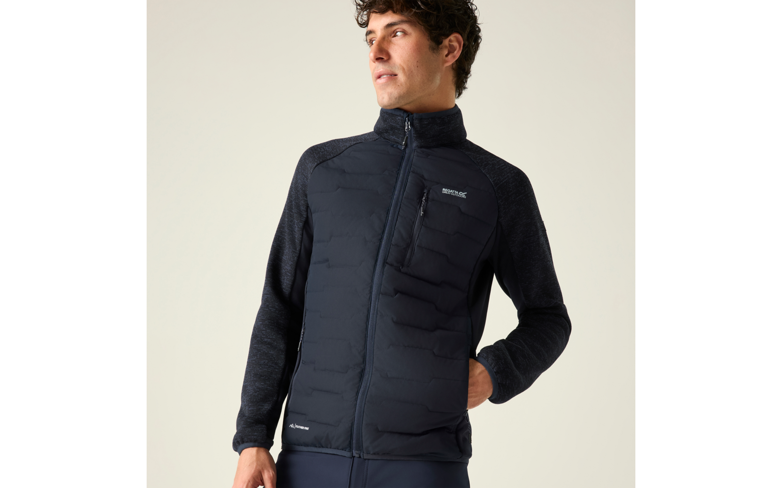 Person mit dunkler gesteppter Jacke mit Frontreißverschluss und sichtbarer Brusttasche, rechte Hand in Seitentasche, Blick nach rechts, heller einfarbiger Hintergrund