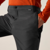 Close-up van taille tot dij van een persoon: donkergrijze broek met knoop, klein merklipje, zijritszak en zichtbare naden; rechterhand in de zak; oranje shirt met lange mouwen
