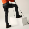Onderlichaam van een persoon met oranje bovenstuk en donkere wandelbroek, zwarte wandelschoen op een wit blok, twee onderarmkrukken, neutrale achtergrond