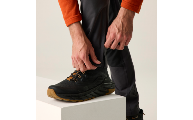 Handen ritsen de pijp van een donkere broek dicht; zwarte schoen met lichtbruine zool op een wit blok; oranje mouw zichtbaar