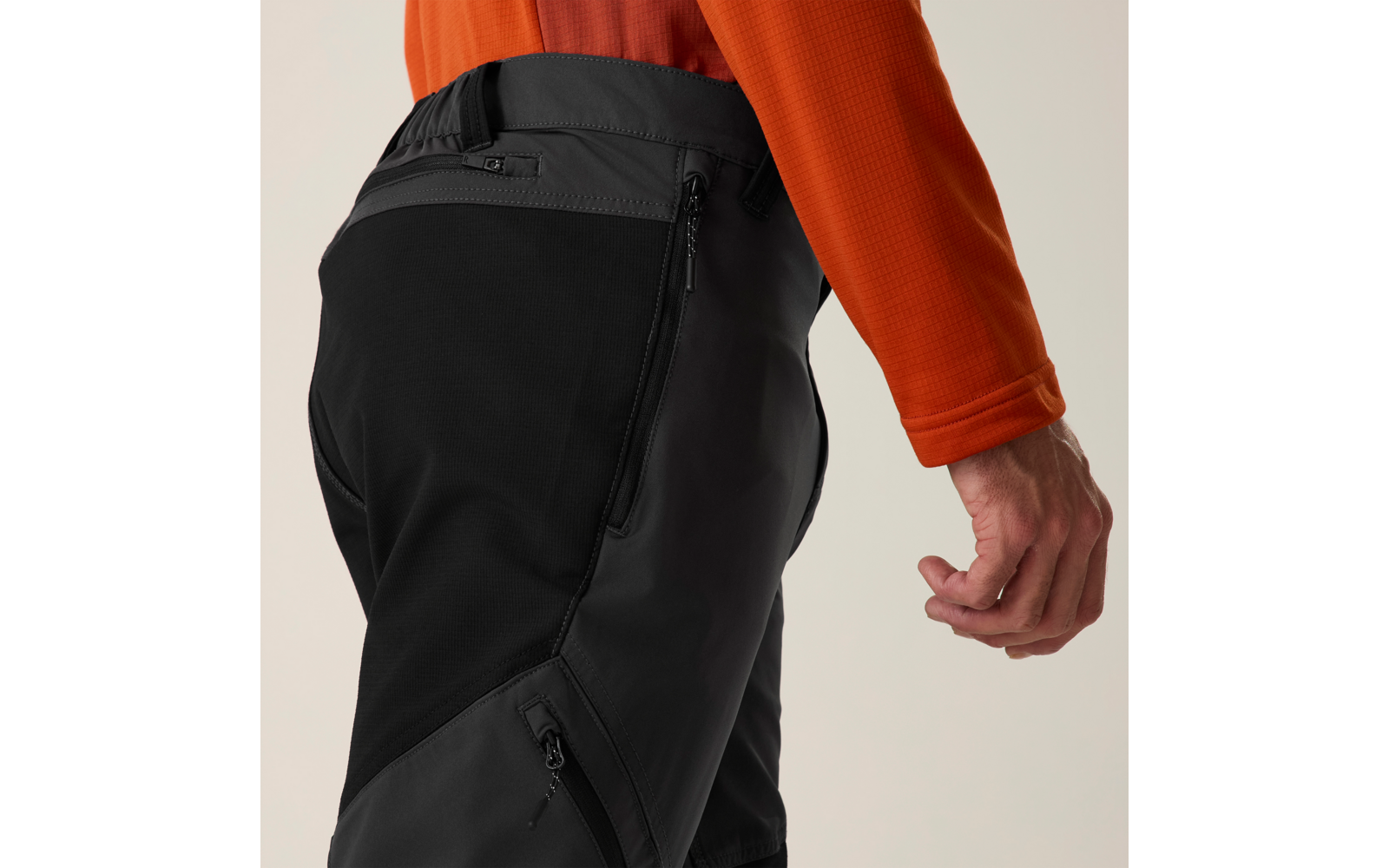 Gedeeltelijke weergave van een persoon van heup tot hand, oranje mouw, zwart-grijze broek met ritszakken