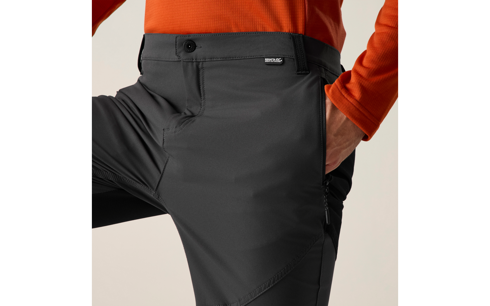 Close-up van taille tot dij van een persoon: donkergrijze broek met knoop, klein merklipje, zijritszak en zichtbare naden; rechterhand in de zak; oranje shirt met lange mouwen