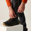 Handen ritsen de pijp van een donkere broek dicht; zwarte schoen met lichtbruine zool op een wit blok; oranje mouw zichtbaar