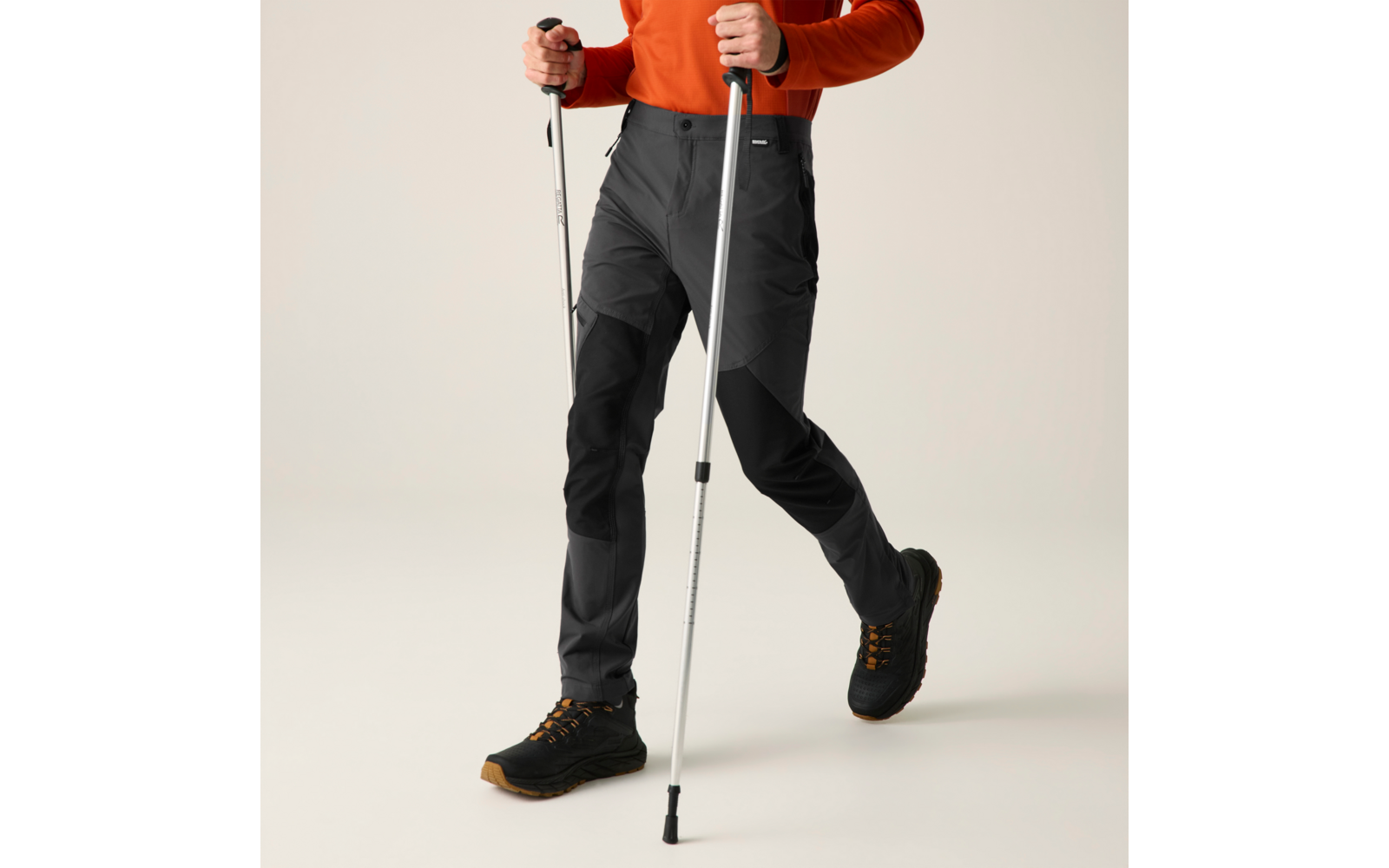 Onderlichaam van een persoon met een oranje bovenstuk, donkergrijze broek, zwarte laarzen met bruine zolen en twee metalen stokken met rubberen uiteinden; één been naar voren, lichte egale achtergrond