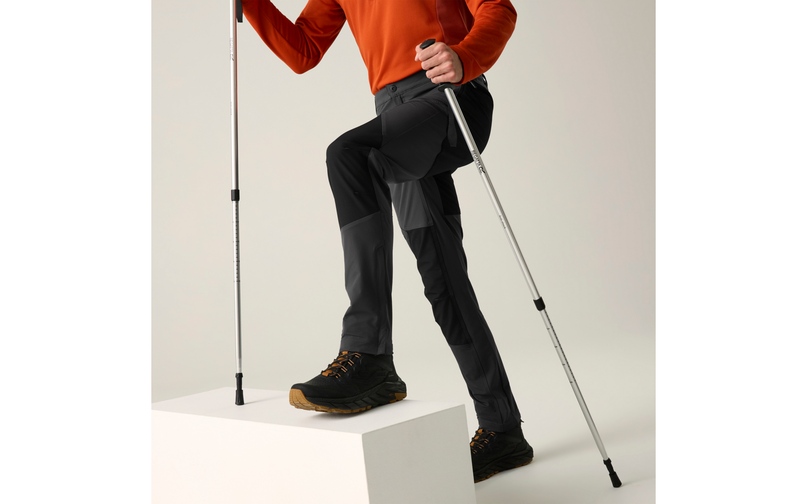Onderlichaam van een persoon in een oranje bovenstuk en zwarte broek, één voet op een wit blok, houdt twee zilverkleurige wandelstokken vast, draagt zwarte wandelschoenen met bruine zolen