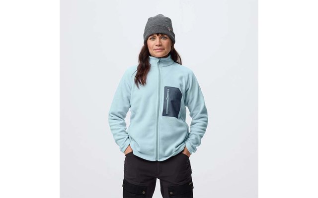 Person vor weißem Hintergrund, trägt hellblaue Fleecejacke mit dunkler Brusttasche, graue Strickmütze, schwarze Cargohose, Hände in den Taschen