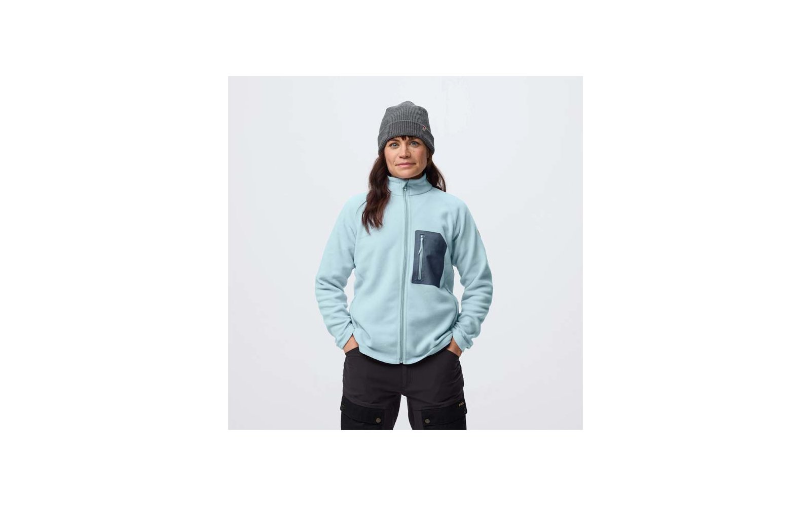 Person vor weißem Hintergrund, trägt hellblaue Fleecejacke mit dunkler Brusttasche, graue Strickmütze, schwarze Cargohose, Hände in den Taschen
