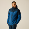 Person in blauer Softshell-Jacke mit Kapuze, steht mit Händen in den Taschen und blickt zur Seite vor hellem, einfarbigem Hintergrund